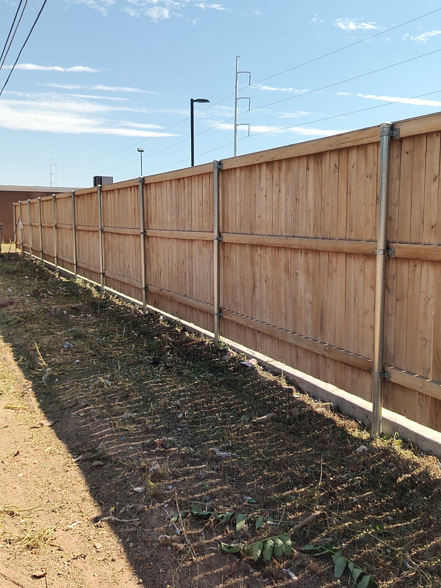 Wooden fence along dirt and grass; utility poles and wires in the background under a blue sky.
