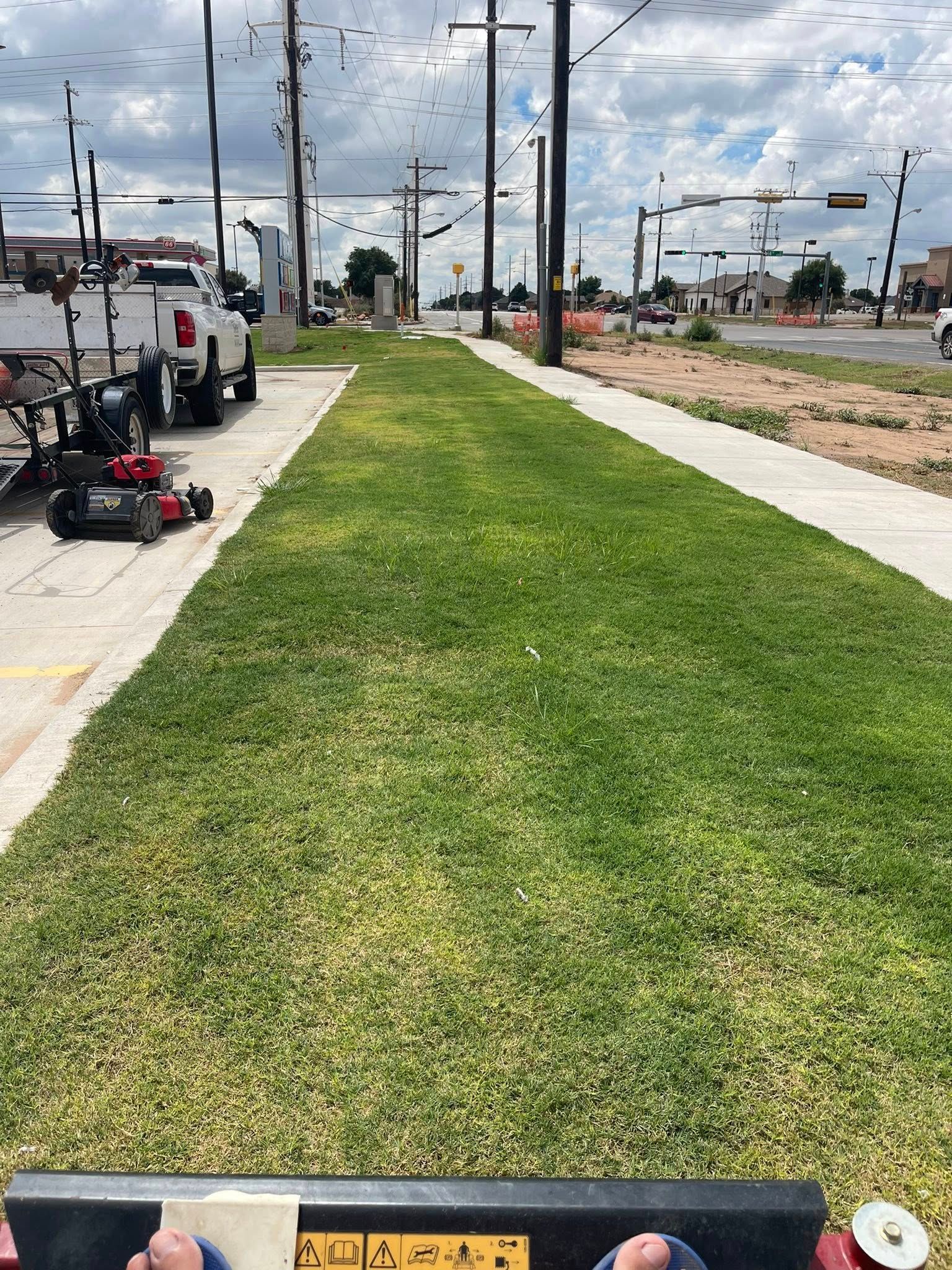 Lawn mowing alongside a sidewalk and road, with a truck and equipment visible. Green grass, blue sky.