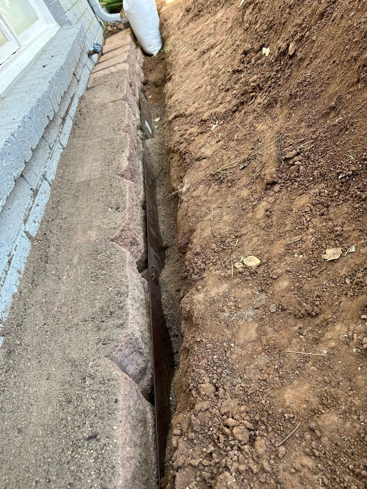 Narrow trench dug next to a brick foundation and building wall, filled with dark soil.