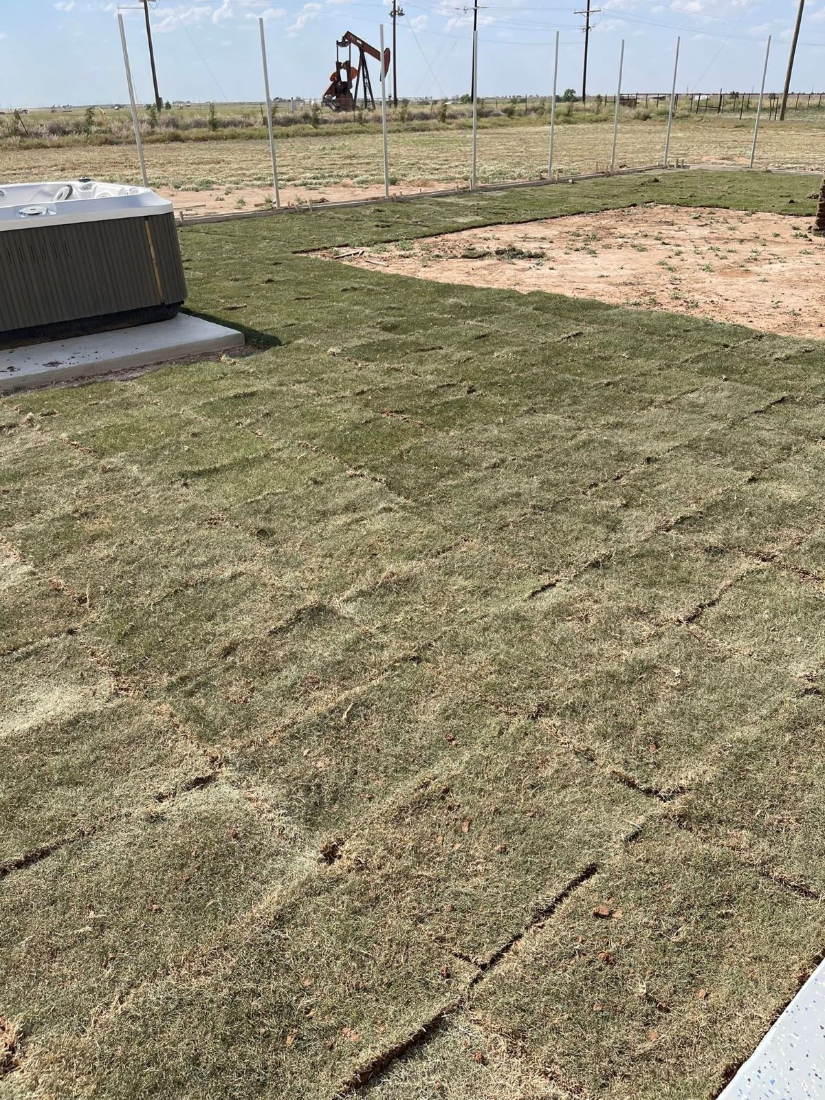 Newly laid sod in a field, with oil pumps visible in the background under a bright sky.