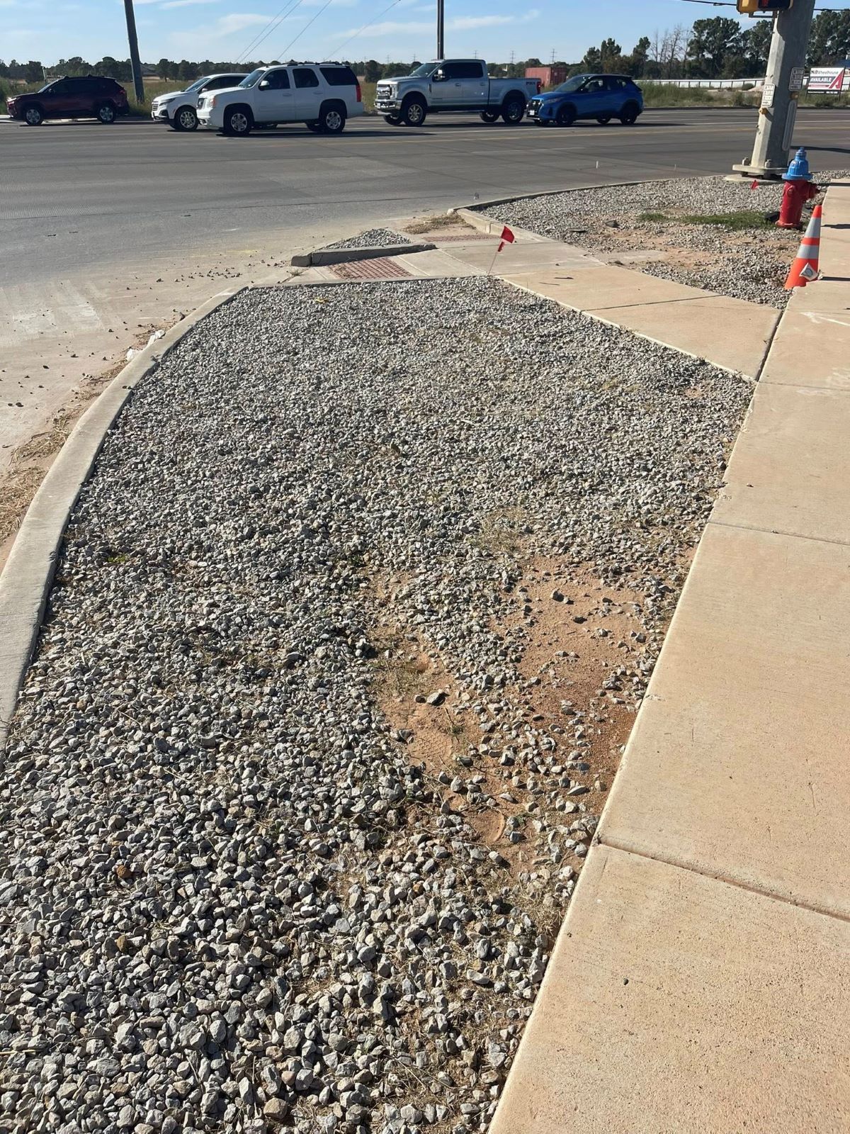 Gravel-filled section beside a sidewalk at a road intersection, with vehicles in the background.
