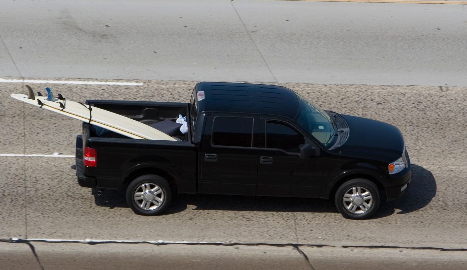 A black truck with a surfboard in the bed is driving down a highway.