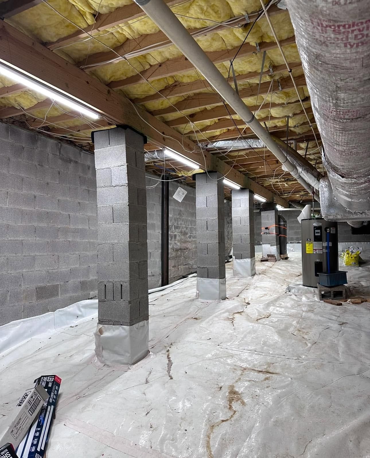 Unfinished basement with cinder block columns, insulation overhead, and white floor covering.