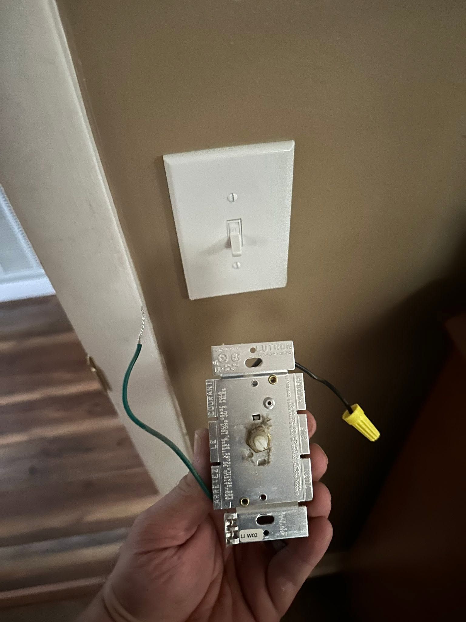 Hand holding a light switch plate with exposed wiring next to a wall outlet on a beige wall
