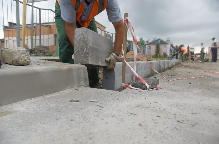A construction worker places a grey concrete curb on a sidewalk, outdoors.