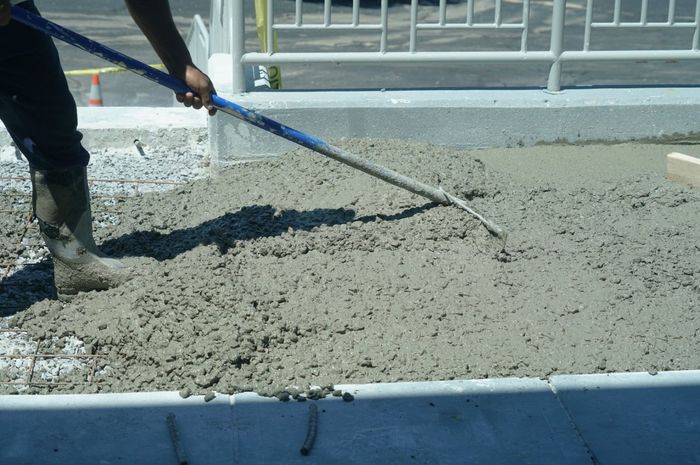 Person using a rake to spread wet concrete on a sidewalk.