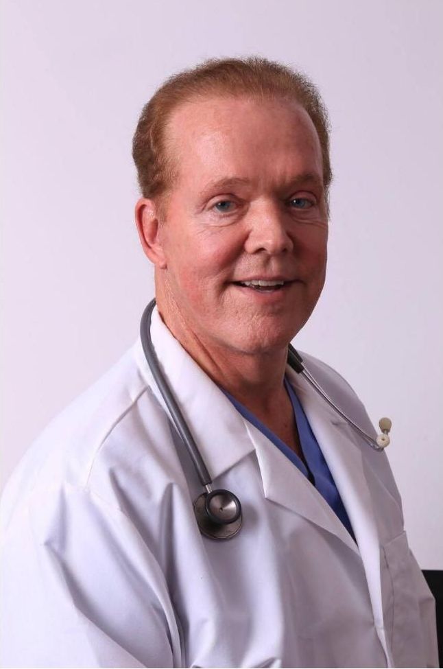 A smiling older doctor with red hair wearing a white coat and stethoscope, against a white background.