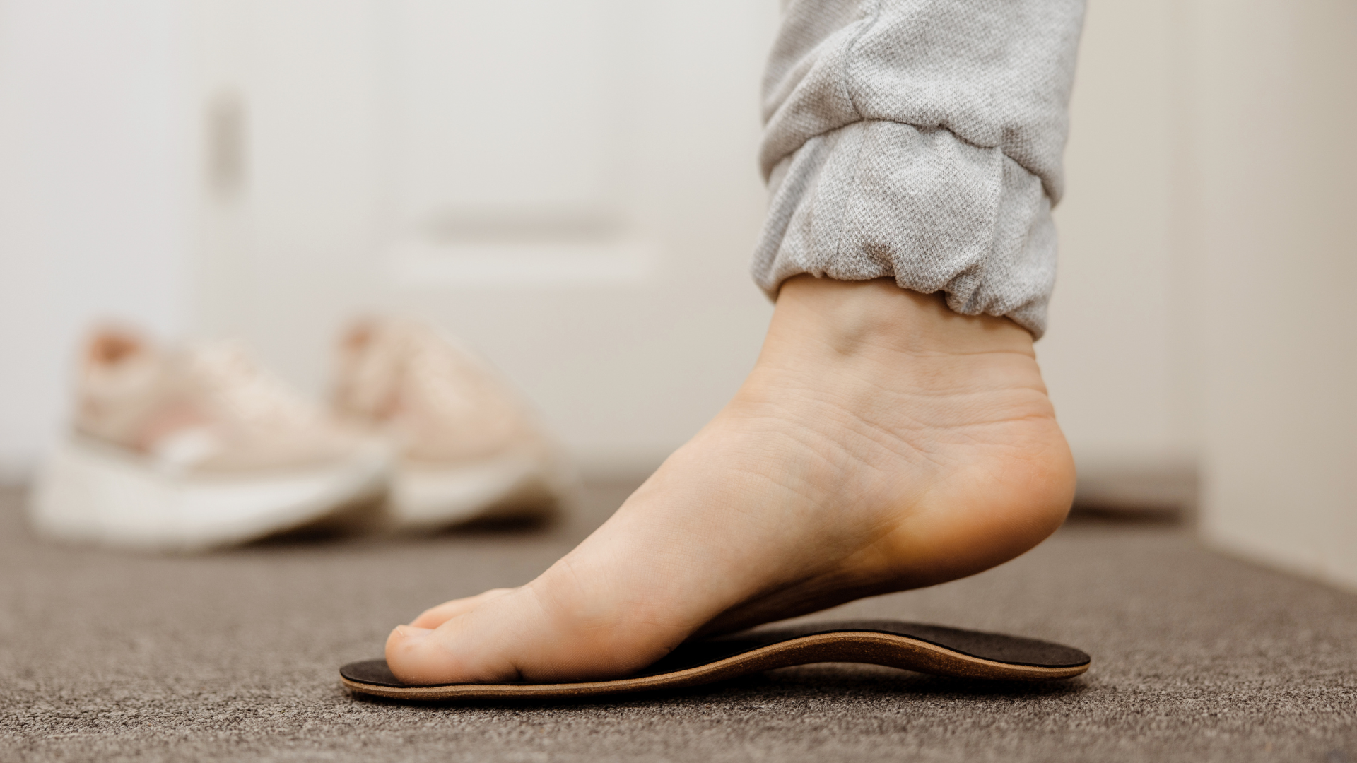 Person's foot wearing a gray sweatpant stands on an orthotic insert. Pink sneakers are in the background.