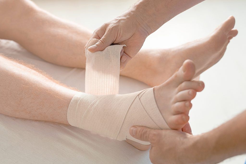 Person's ankle being bandaged. Tan skin, white bandage being wrapped around the ankle by two hands.