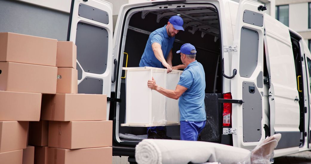 Two workers in blue uniforms loading a white cabinet into the back of a moving van filled with cardboard boxes.