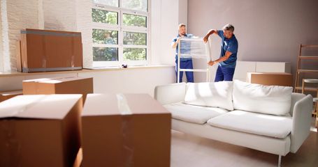 Two movers in blue uniforms move a wrapped furniture item in a room filled with cardboard boxes and a white sofa.