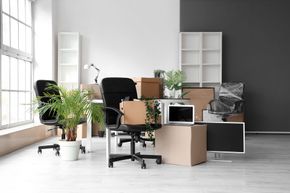 An office during a move, featuring two black chairs, cardboard packing boxes, a computer monitor, and potted plants.