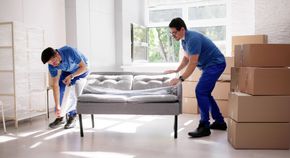 Two movers in blue uniforms wrap a gray sofa in plastic inside a room with stacked cardboard moving boxes.