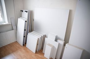 Unassembled white furniture panels stacked against a wall in a room with light wood flooring.