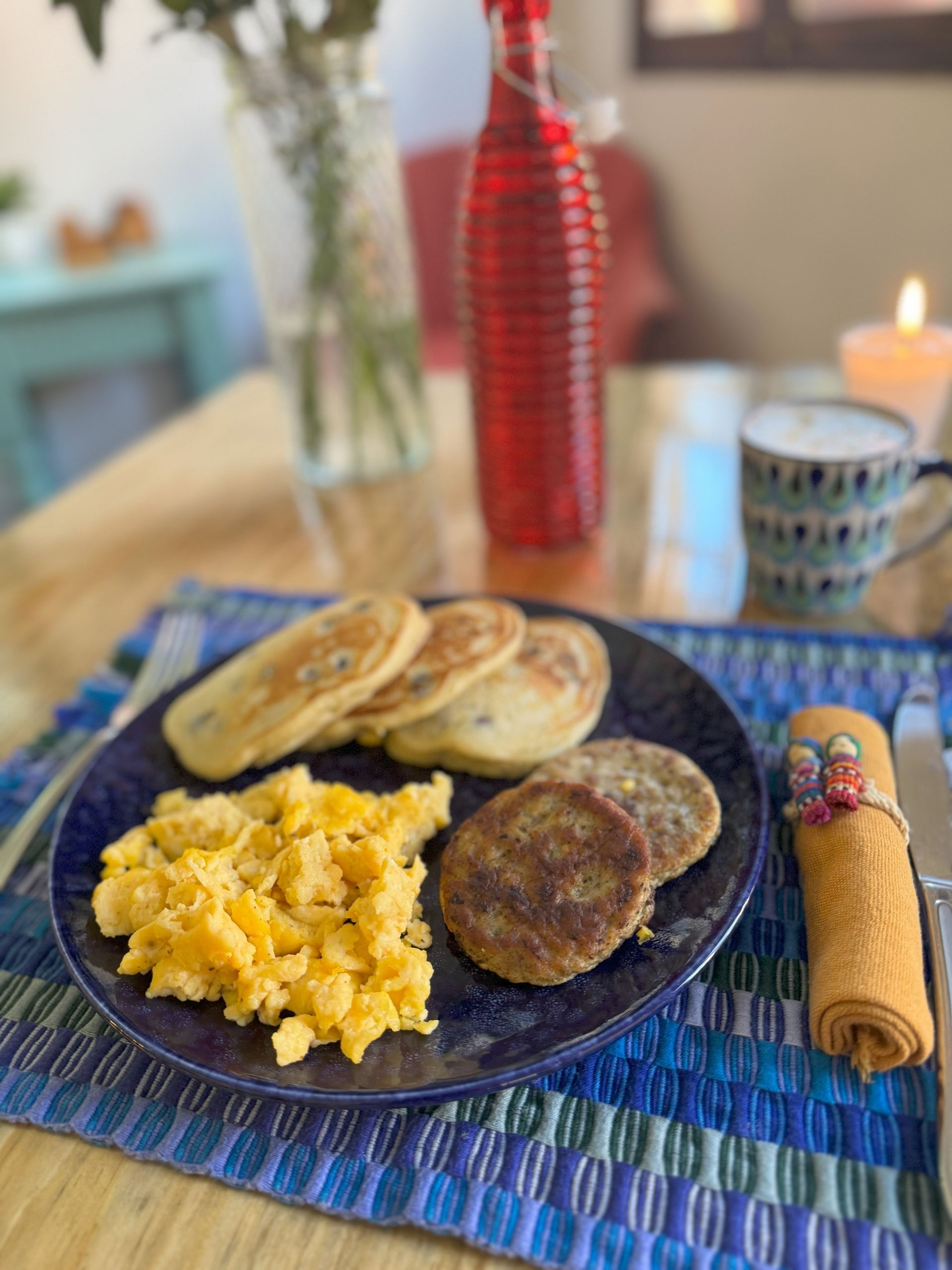 A plate of food with pancakes , scrambled eggs and sausages on a table.