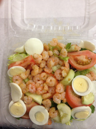 Shrimp salad in a clear plastic container with lettuce, tomatoes, cucumbers, and boiled eggs.