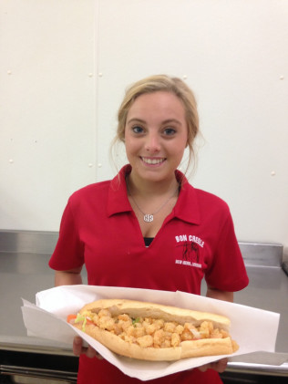 Woman in red shirt smiling, holding a shrimp po'boy sandwich. Light background.
