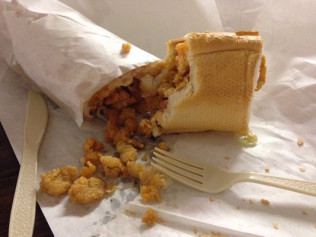 Sub sandwich with fried food spilling out of the bread, on a paper wrapper.