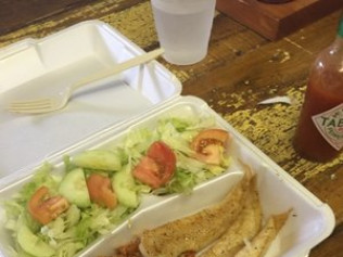 Lunchbox with salad, fish, plastic fork, water glass, and Tabasco sauce on a wooden table.