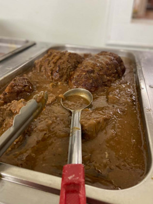 A tray of brown pot roast in gravy with a serving spoon and tongs.