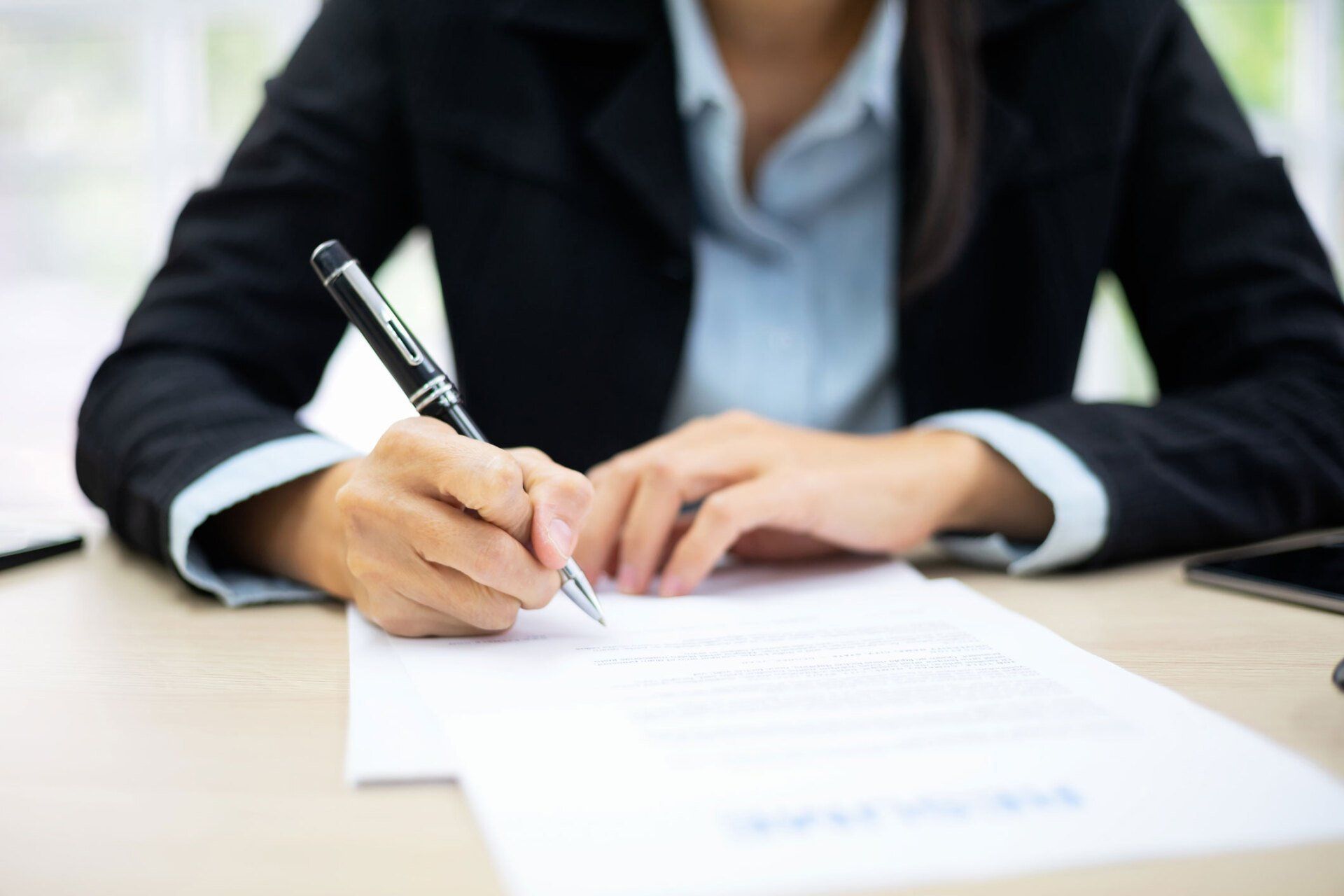 Woman in black blazer writing on a document with a pen at a desk. Woman in black blazer writing on a document with a pen at a desk.