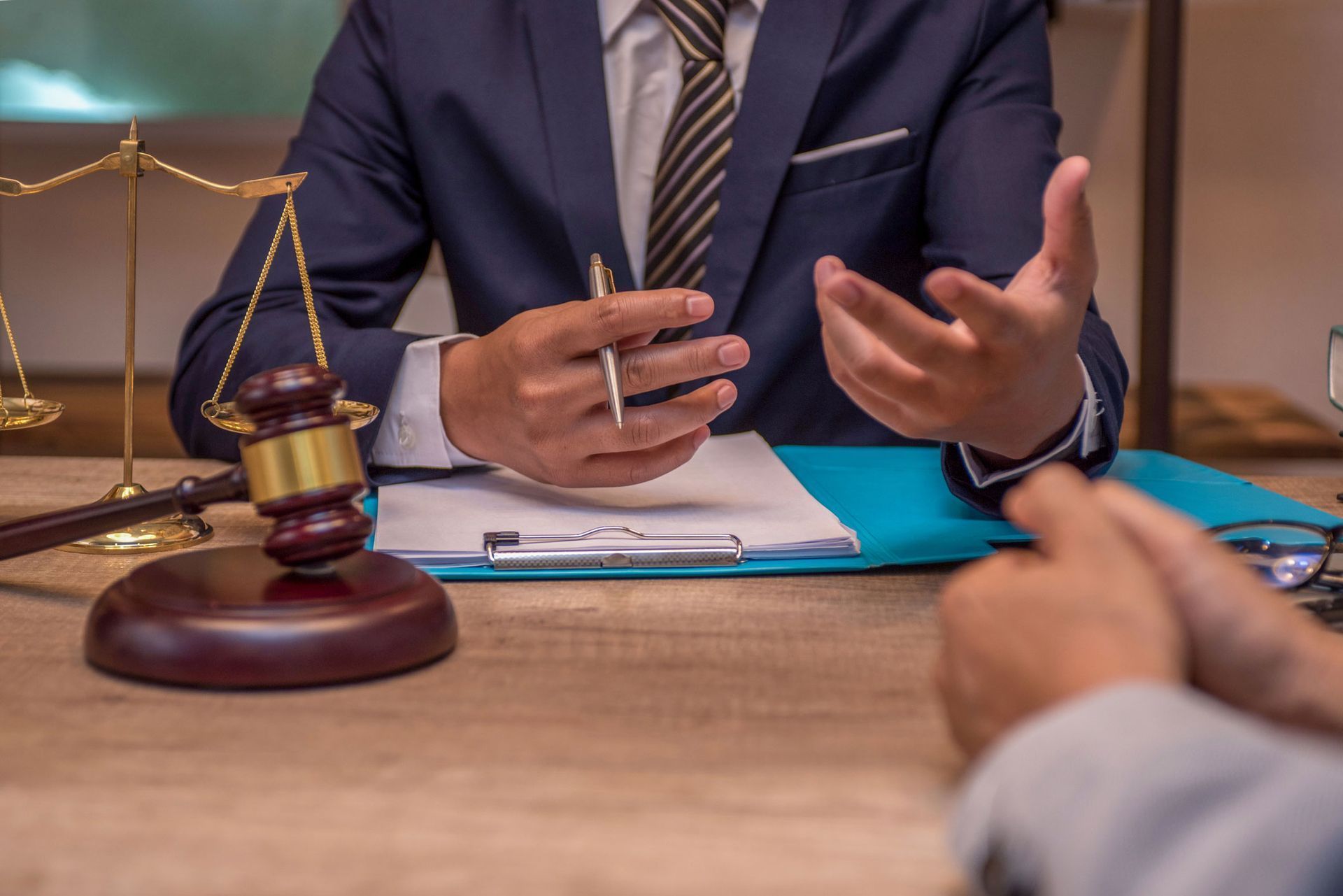 Lawyer in suit gestures, discussing paperwork with a client at a wooden desk with scales and gavel. Lawyer in suit gestures, discussing paperwork with a client at a wooden desk with scales and gavel.