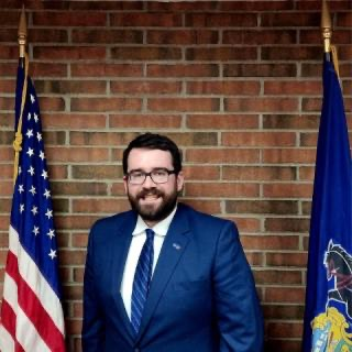 Man in blue suit, smiling, standing between US and state flags, brick wall background. Man in blue suit, smiling, standing between US and state flags, brick wall background.