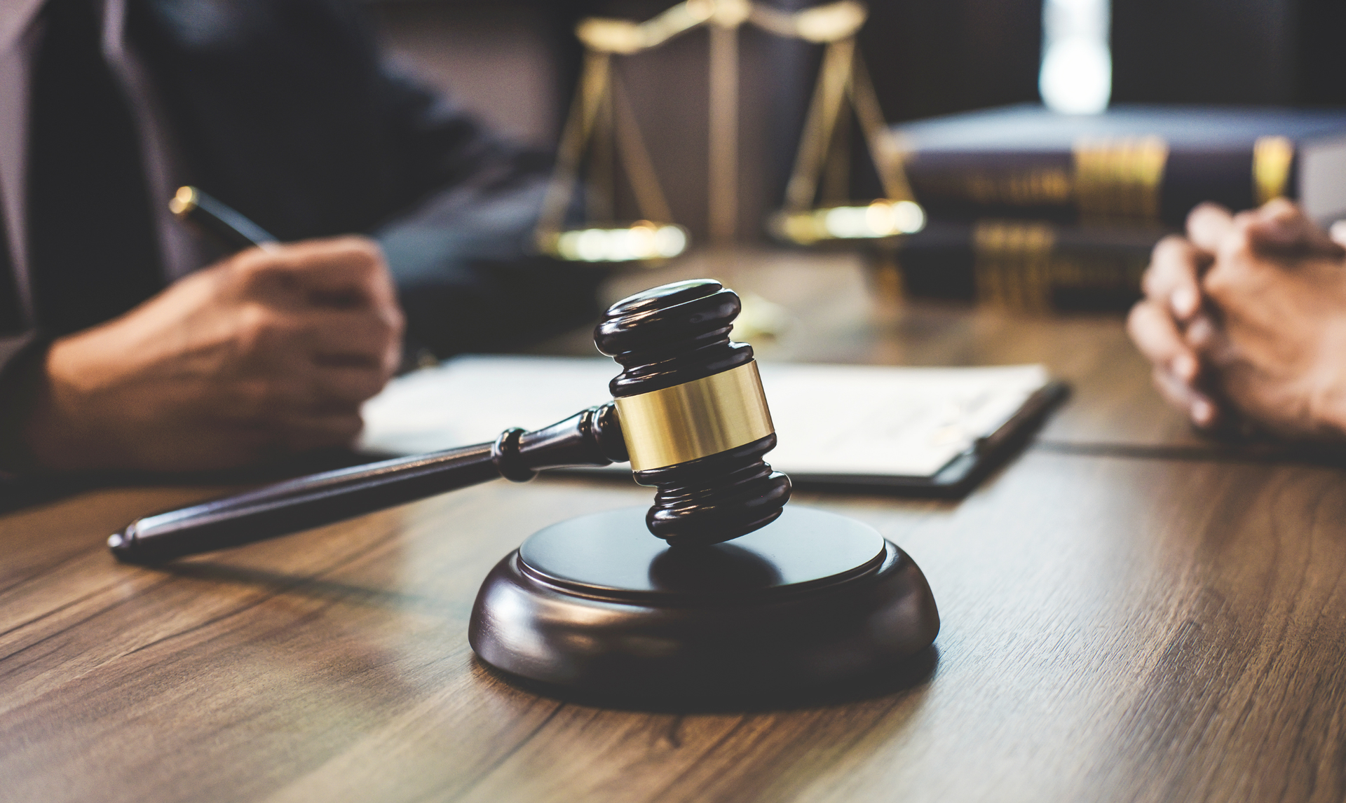 Gavel on a wooden desk, scales of justice in background, hands holding a pen and paperwork.