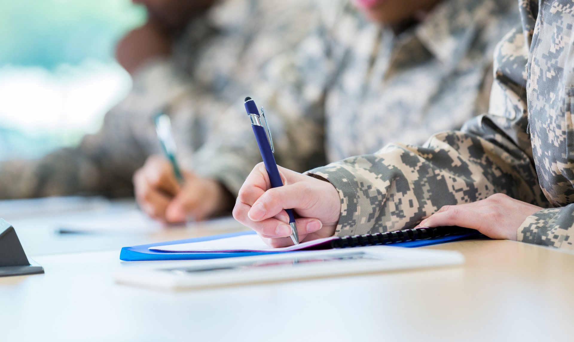 People in camouflage uniforms writing on paper at a table.