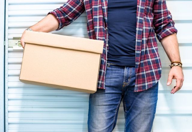 Person holding cardboard box in front of a storage unit door, wearing jeans and a plaid shirt.