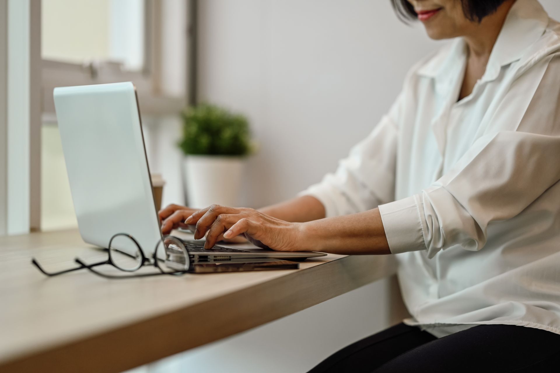 Person typing on a laptop at a desk beside glasses and a small white monitor in a bright workspace
