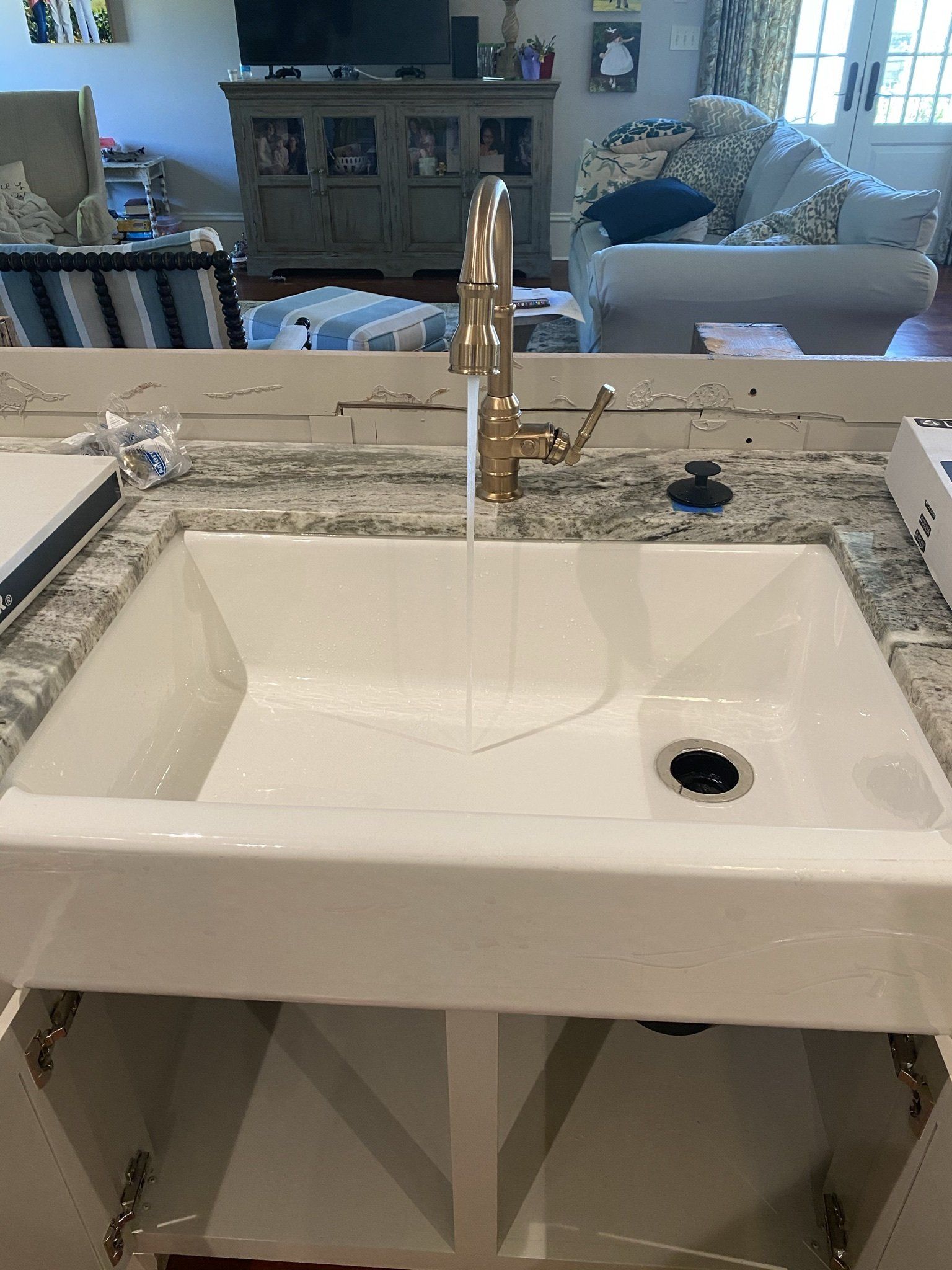 A white kitchen sink with a gold faucet and a living room in the background.