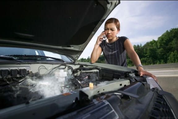 A woman is talking on a cell phone while looking under the hood of a broken down car.