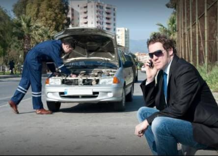 A man is talking on a cell phone while another man works on a car.