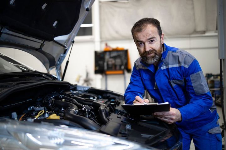 A man is standing in front of a car with the hood open and writing on a clipboard.