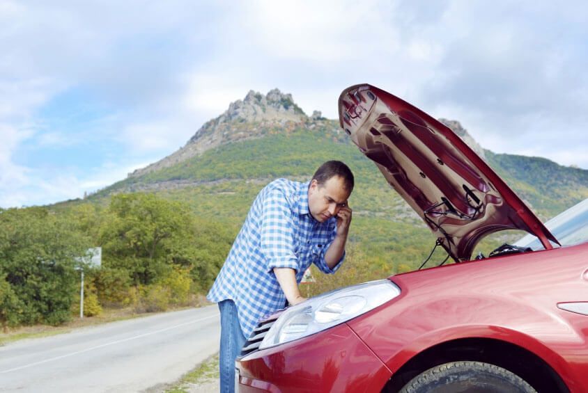 A man is looking under the hood of a broken down car while talking on a cell phone.