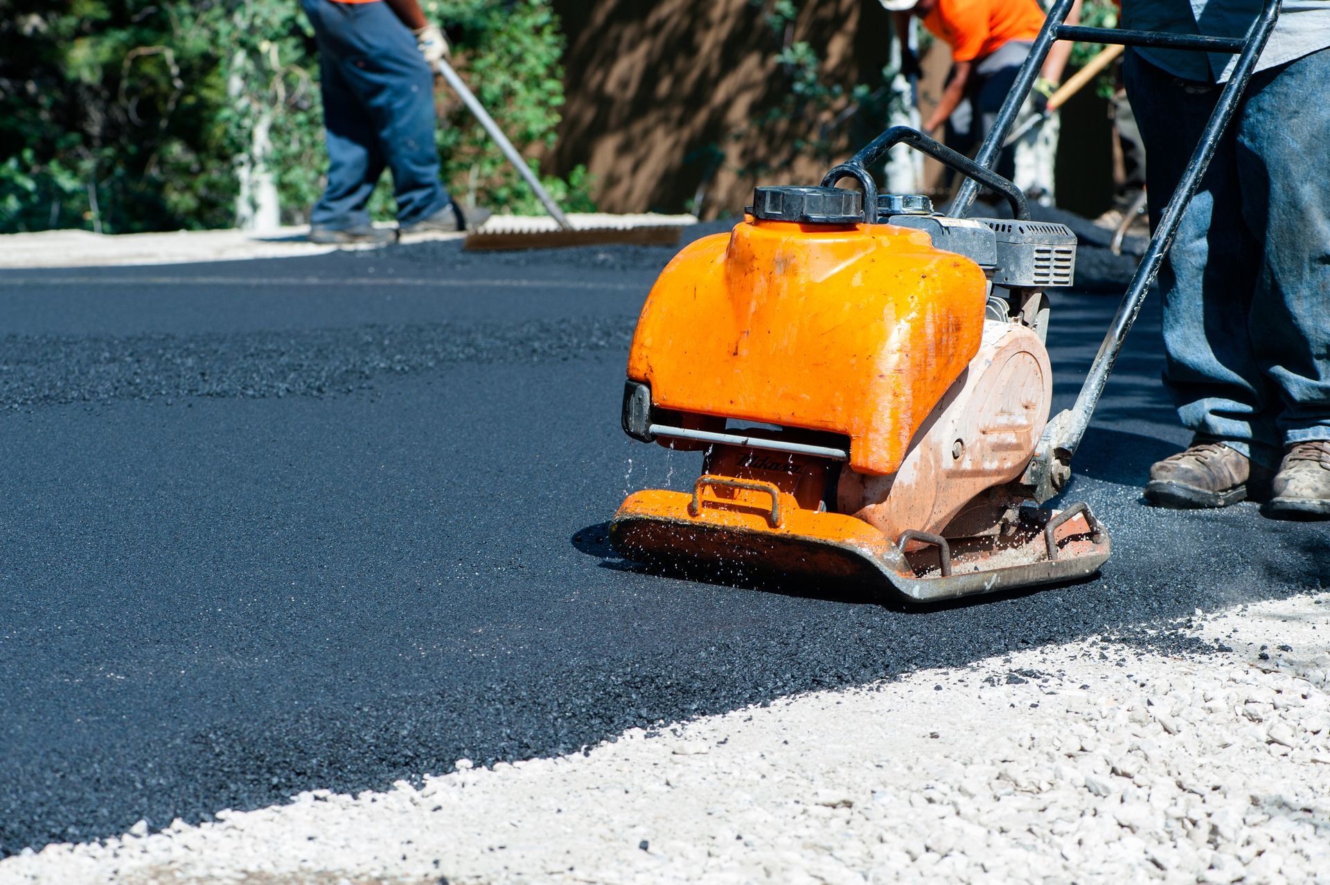 Small orange compactor is rolling asphalt paving on road, with people working on background.