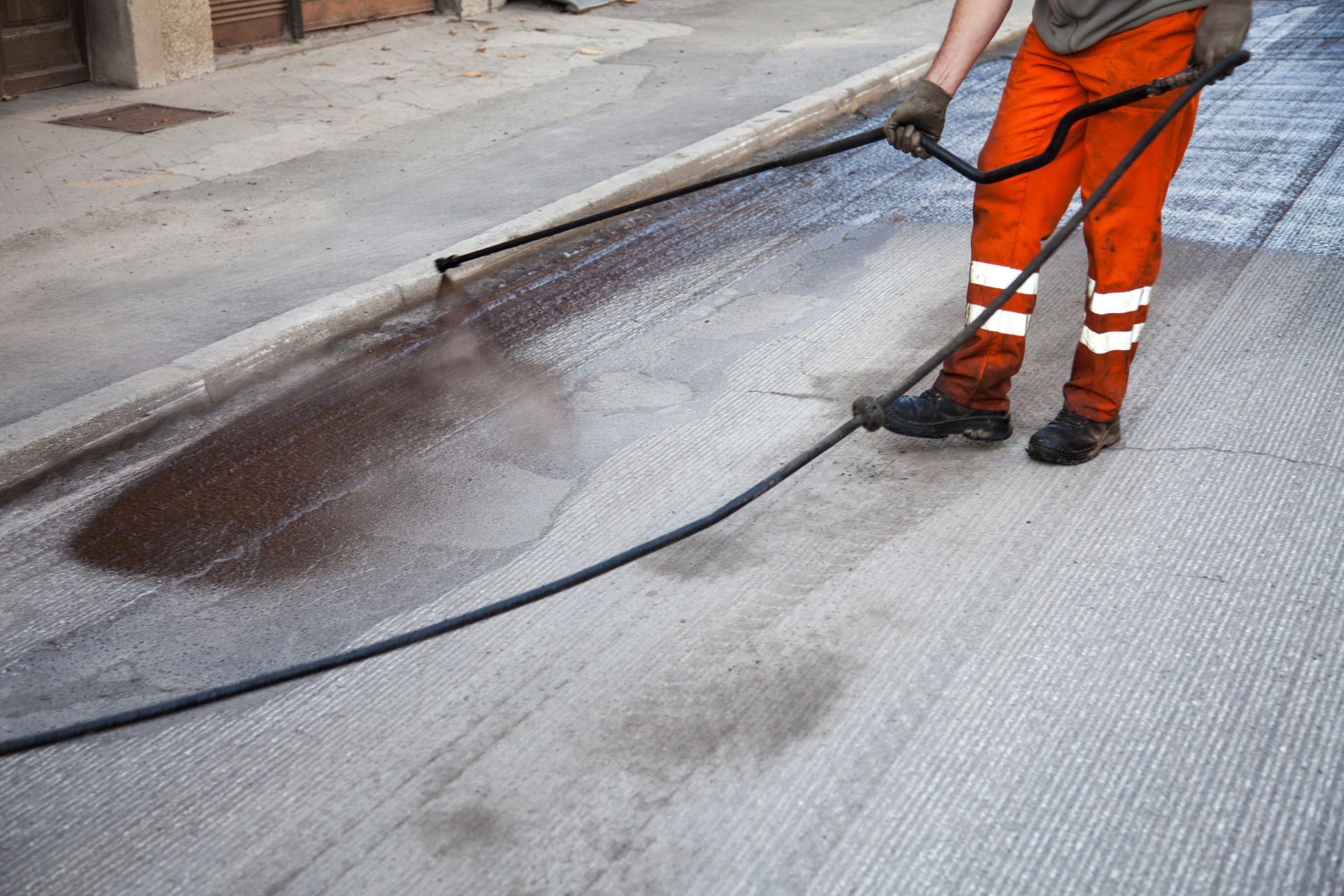Worker spraying sealcoat on road surface, creating a dark, wet layer over dry pavement.
