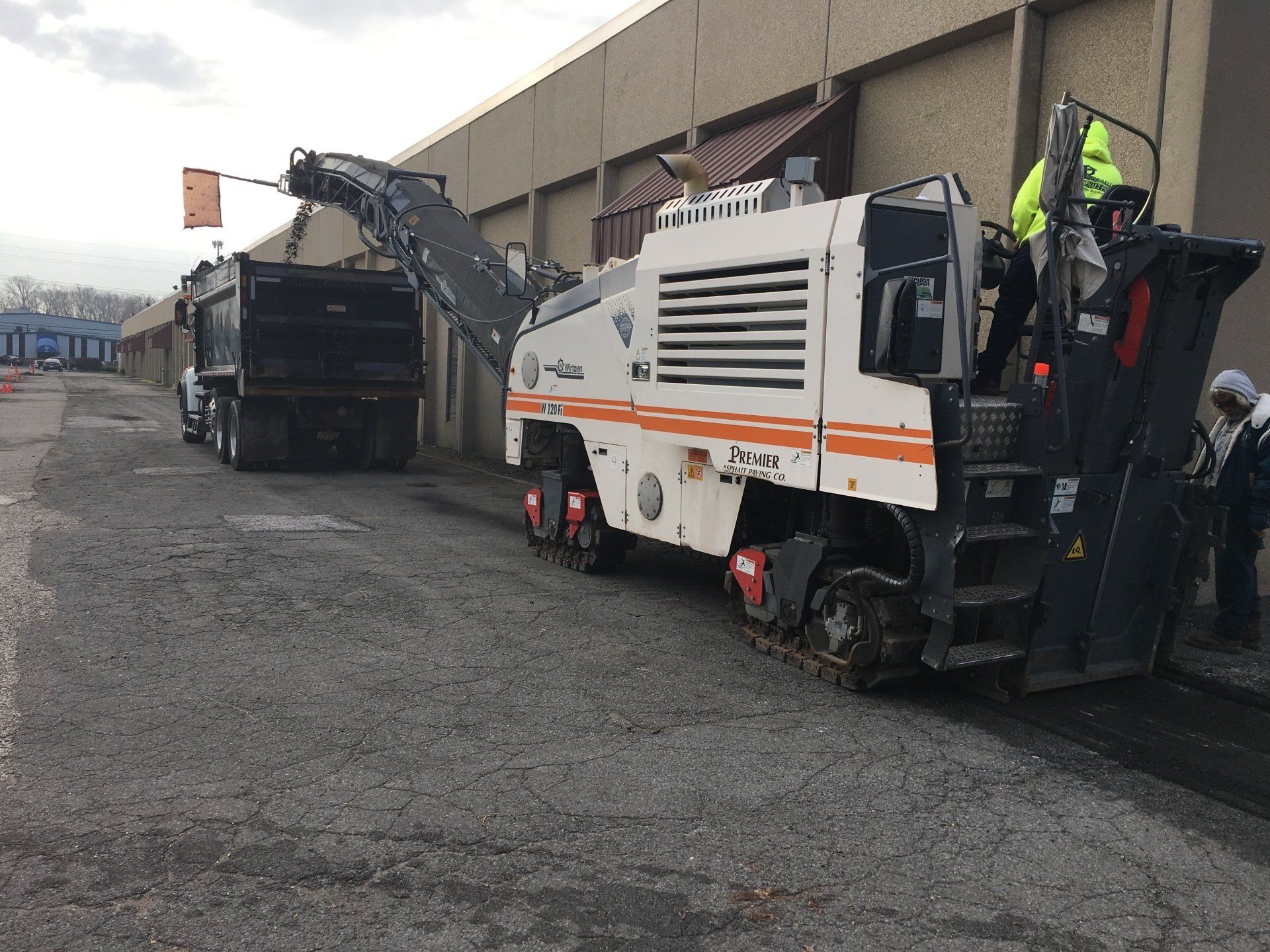 Asphalt milling machine loading truck on a gray road next to a building.