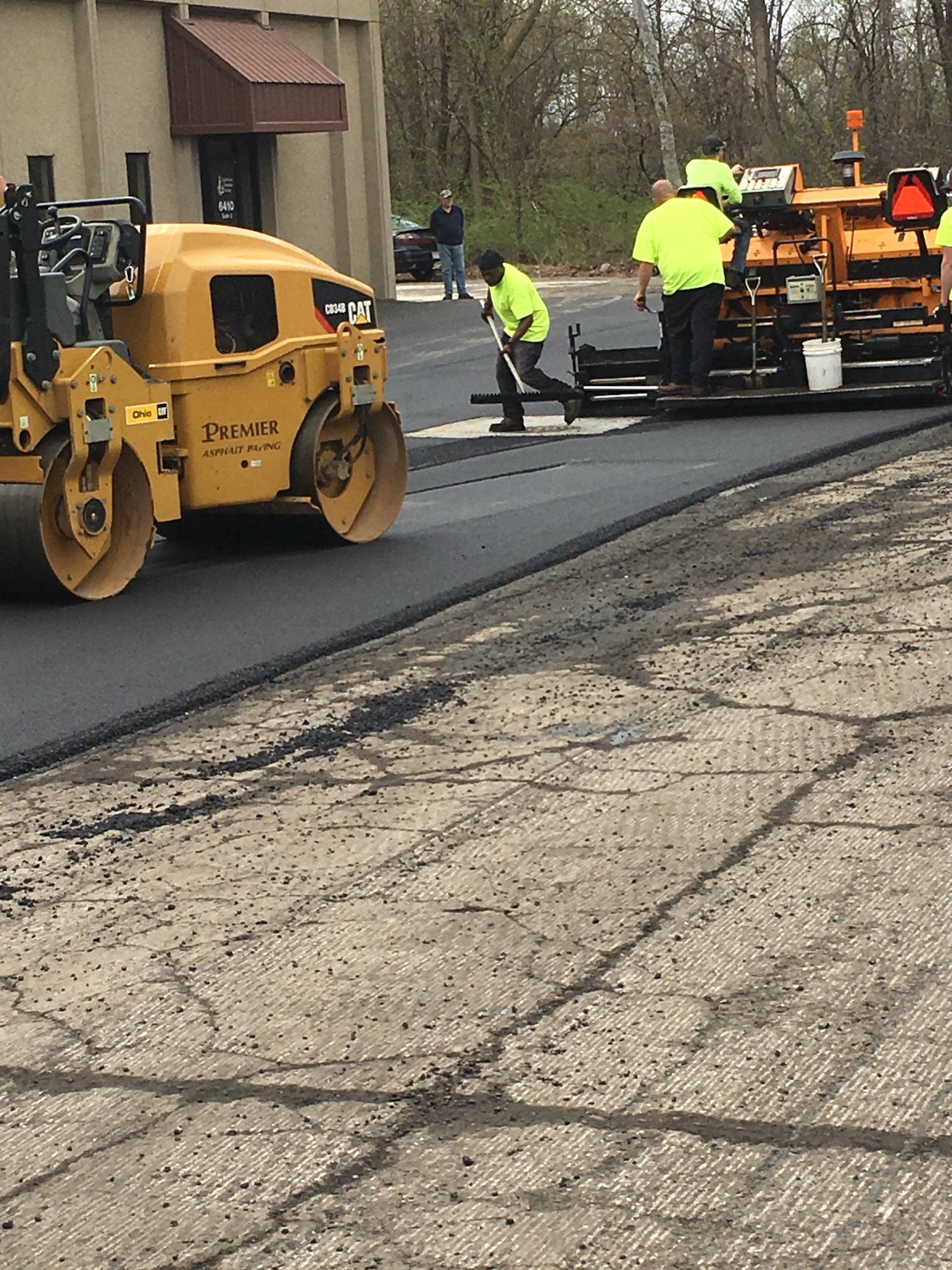 Road paving in progress: Asphalt roller, paver, workers in safety vests smoothing fresh blacktop.