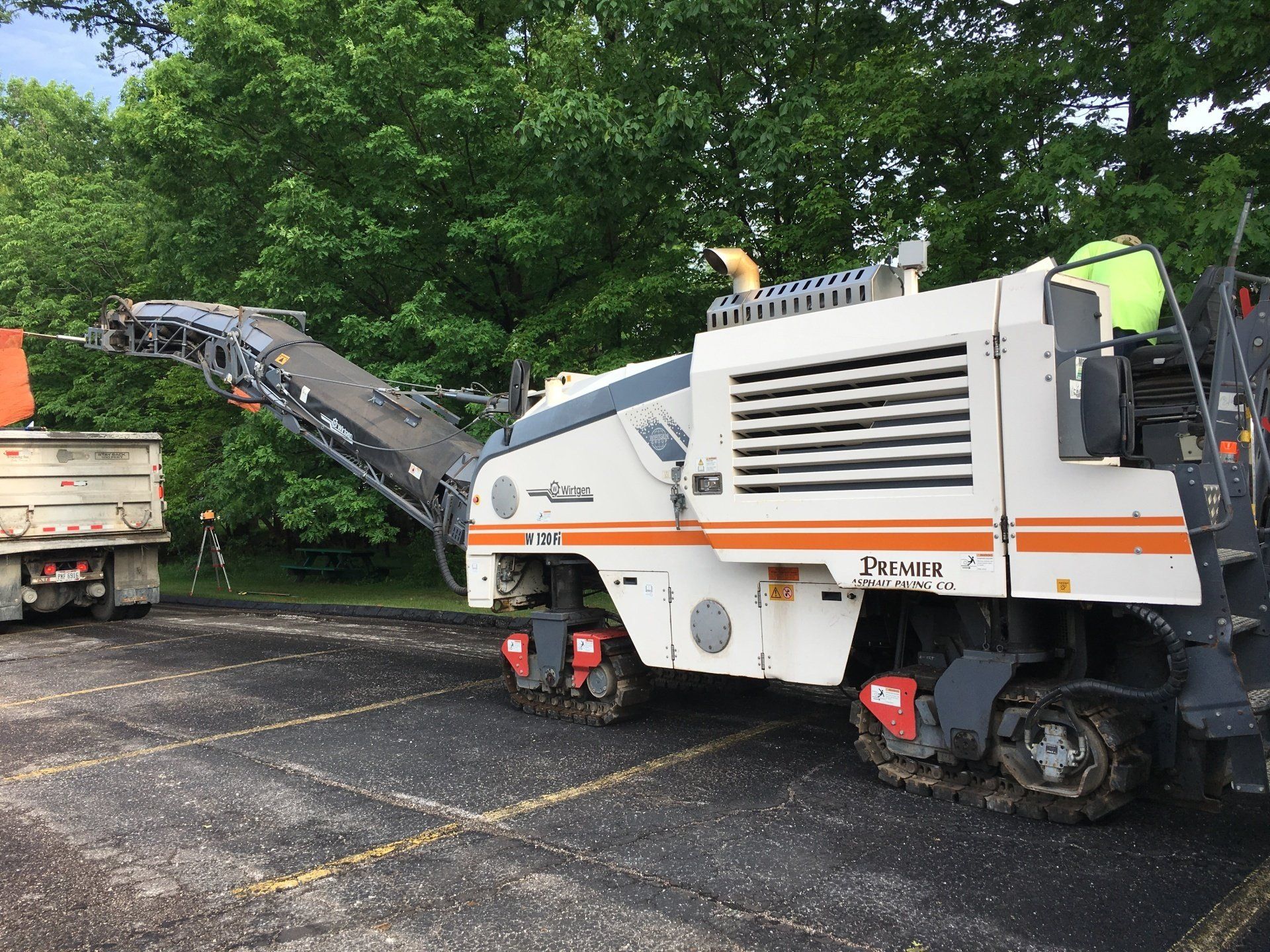 A white asphalt milling machine on a road, removing asphalt. A dump truck is nearby.
