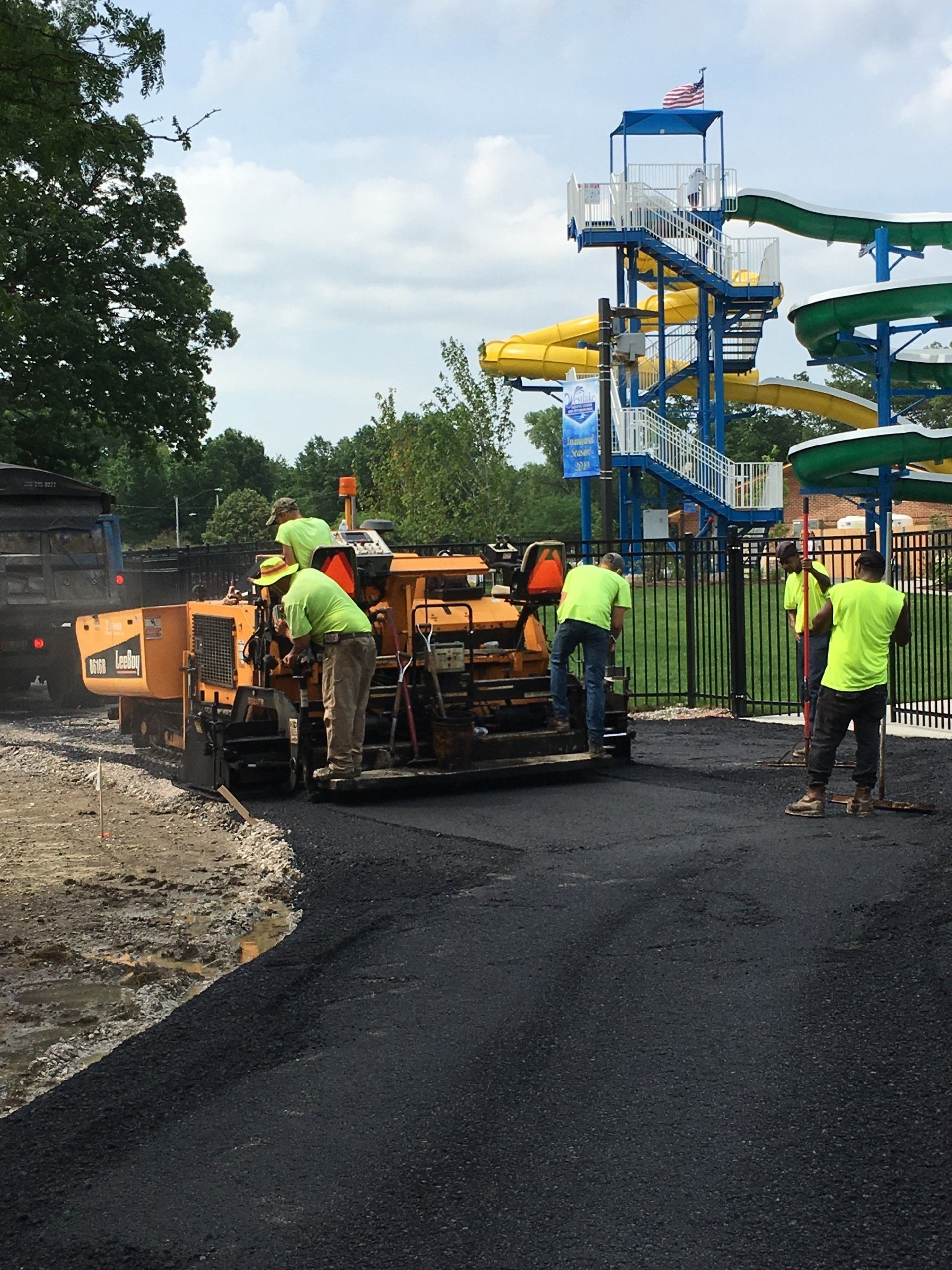 Construction crew paving a road near a waterpark with yellow slides; workers wearing green vests.