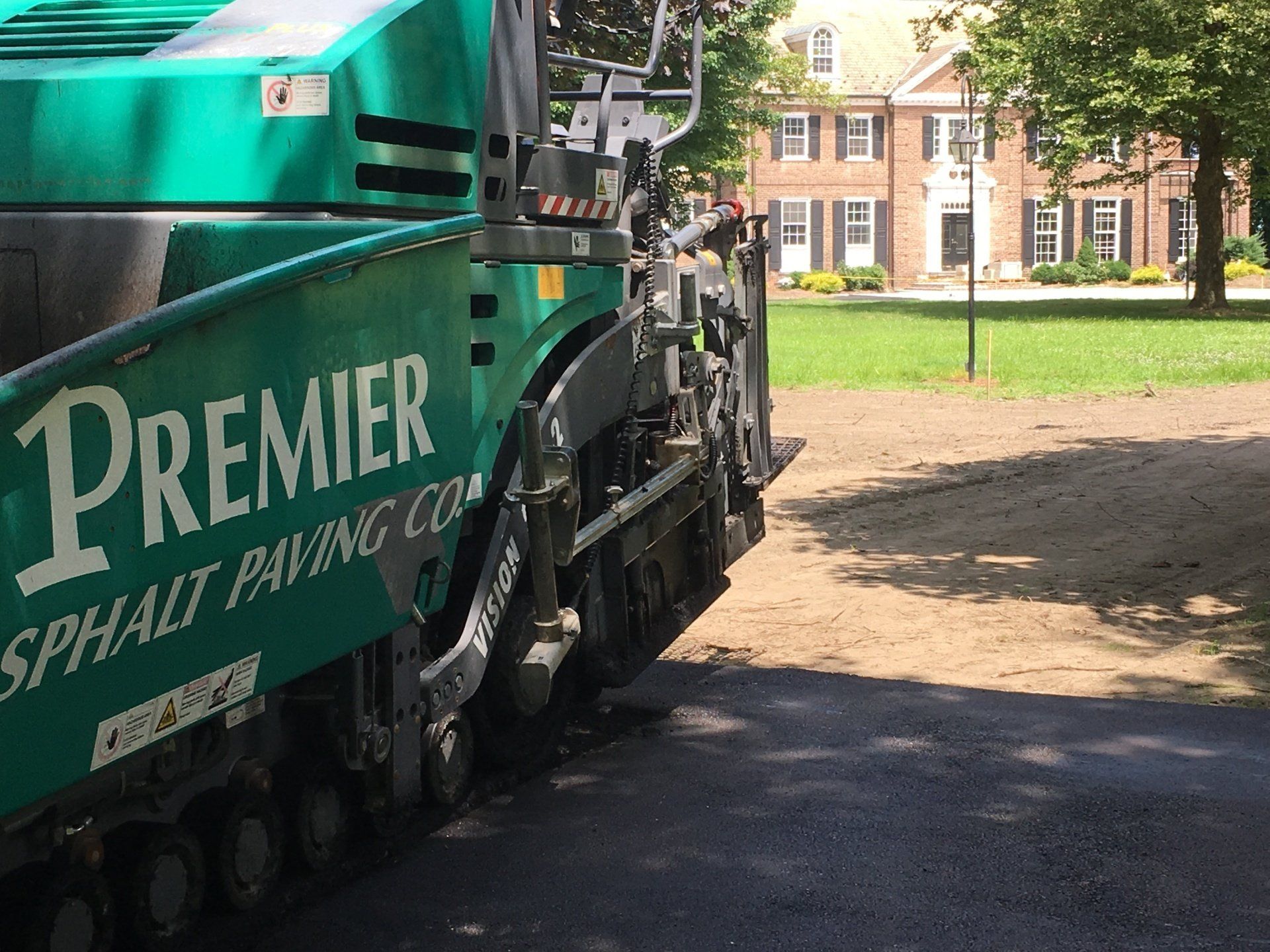 Green asphalt paving machine laying pavement, Premier Asphalt Paving Co, in front of a brick house.
