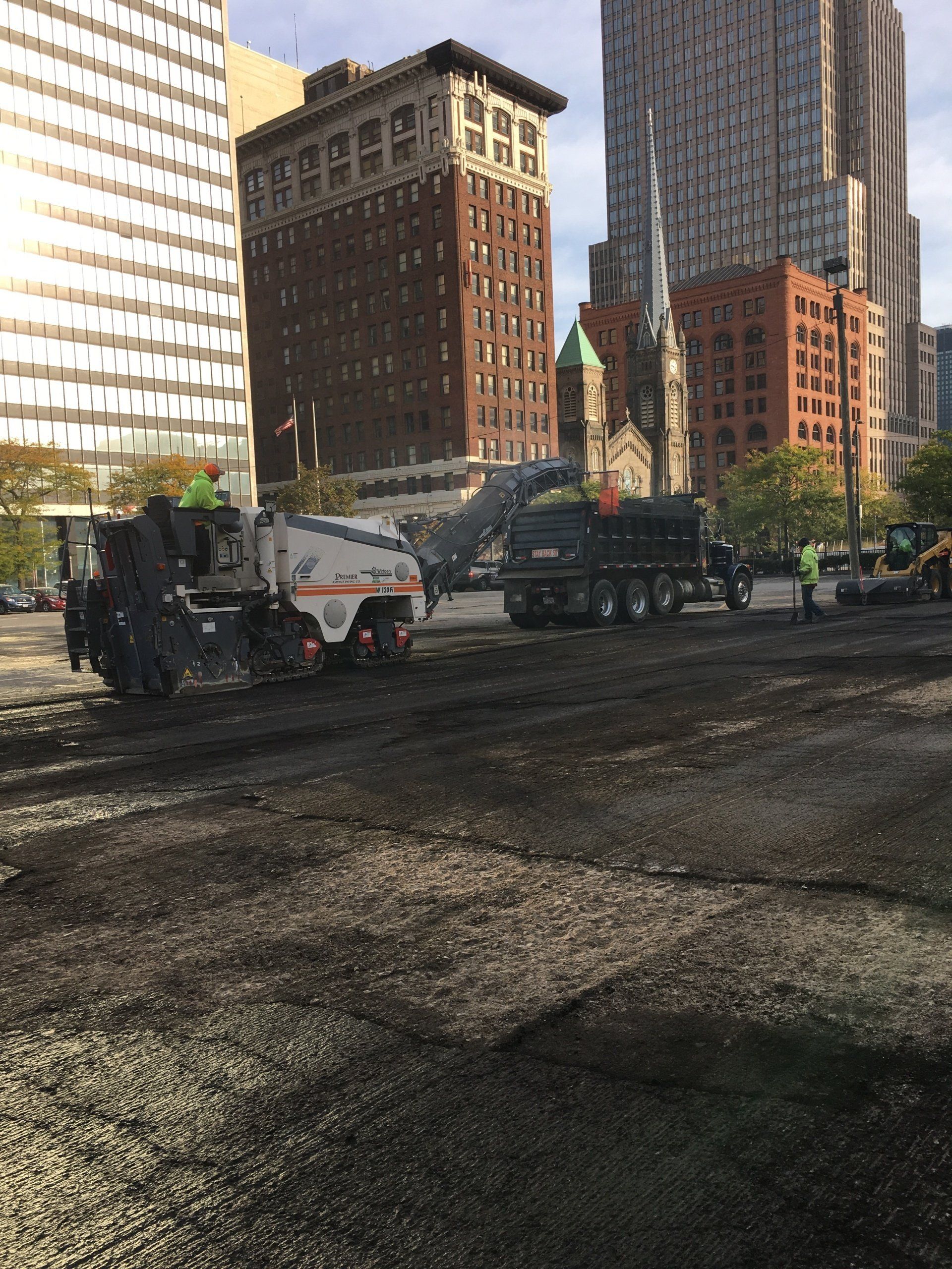Road milling machine removing asphalt; downtown with skyscrapers in background.