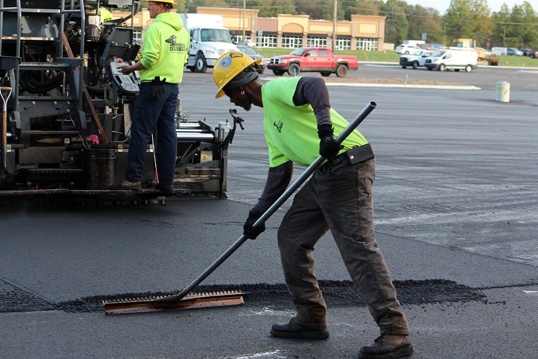 Man in high-vis shirt and hard hat smoothing asphalt with a rake during road work.