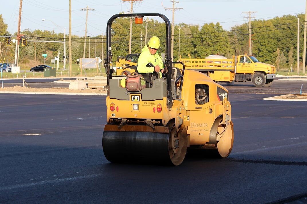 Yellow road roller compacting asphalt with worker wearing a yellow safety helmet.