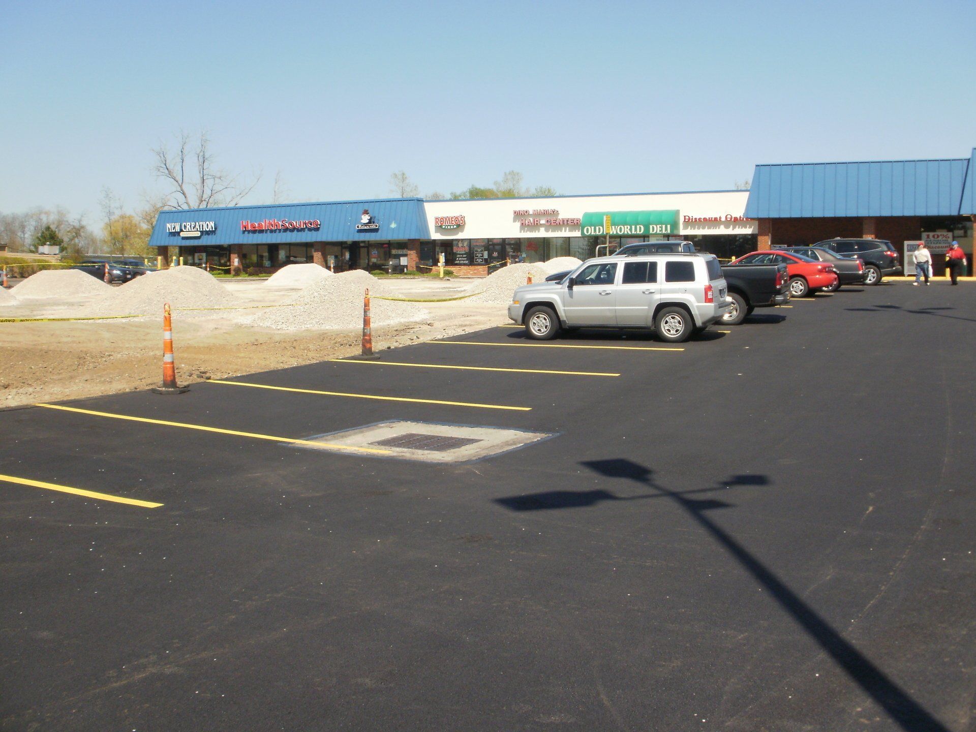 Newly paved parking lot in front of a strip mall with several cars parked. Construction materials in the distance.