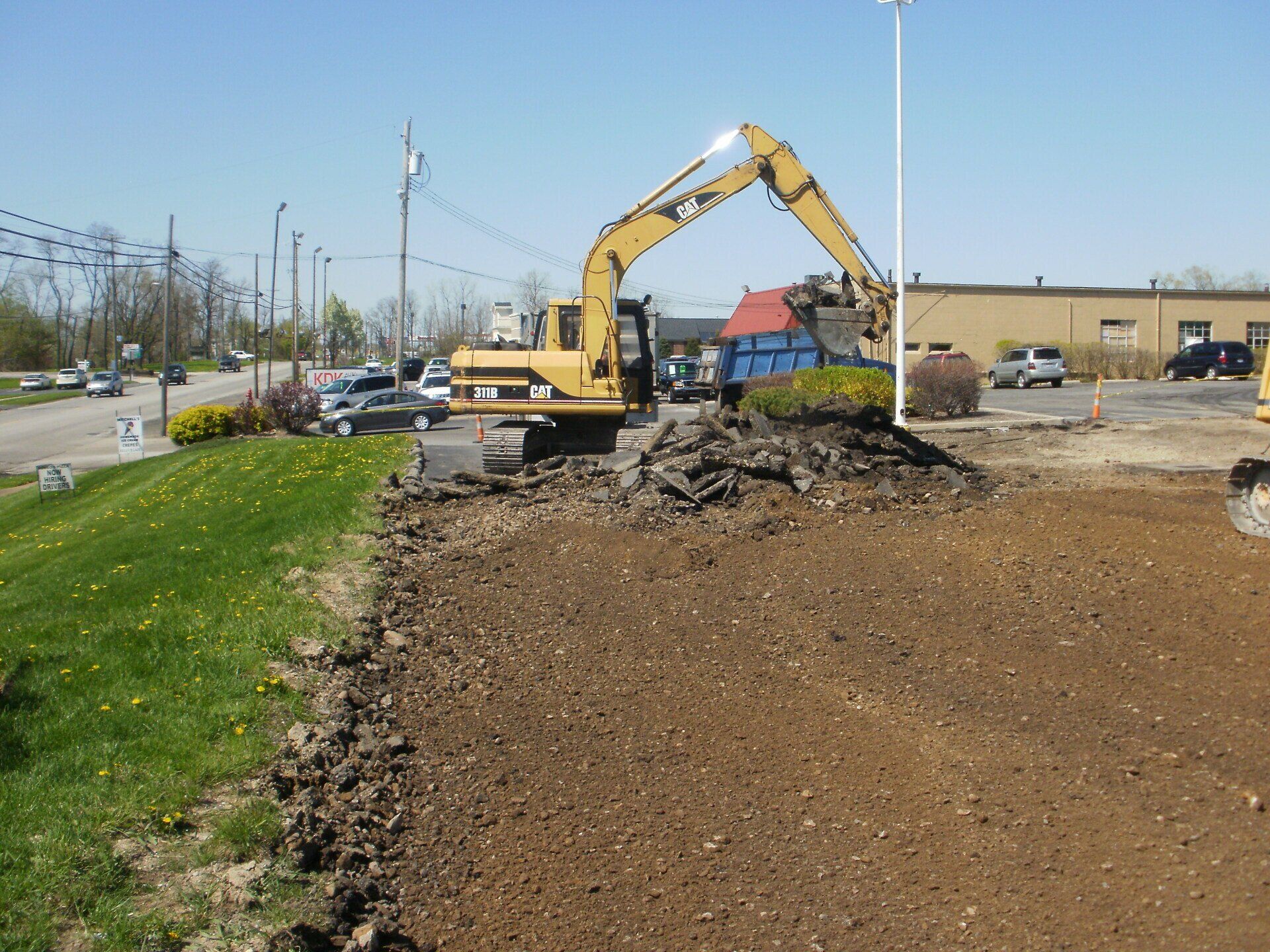 A yellow excavator tears up asphalt on a sunny day beside grass and a road.