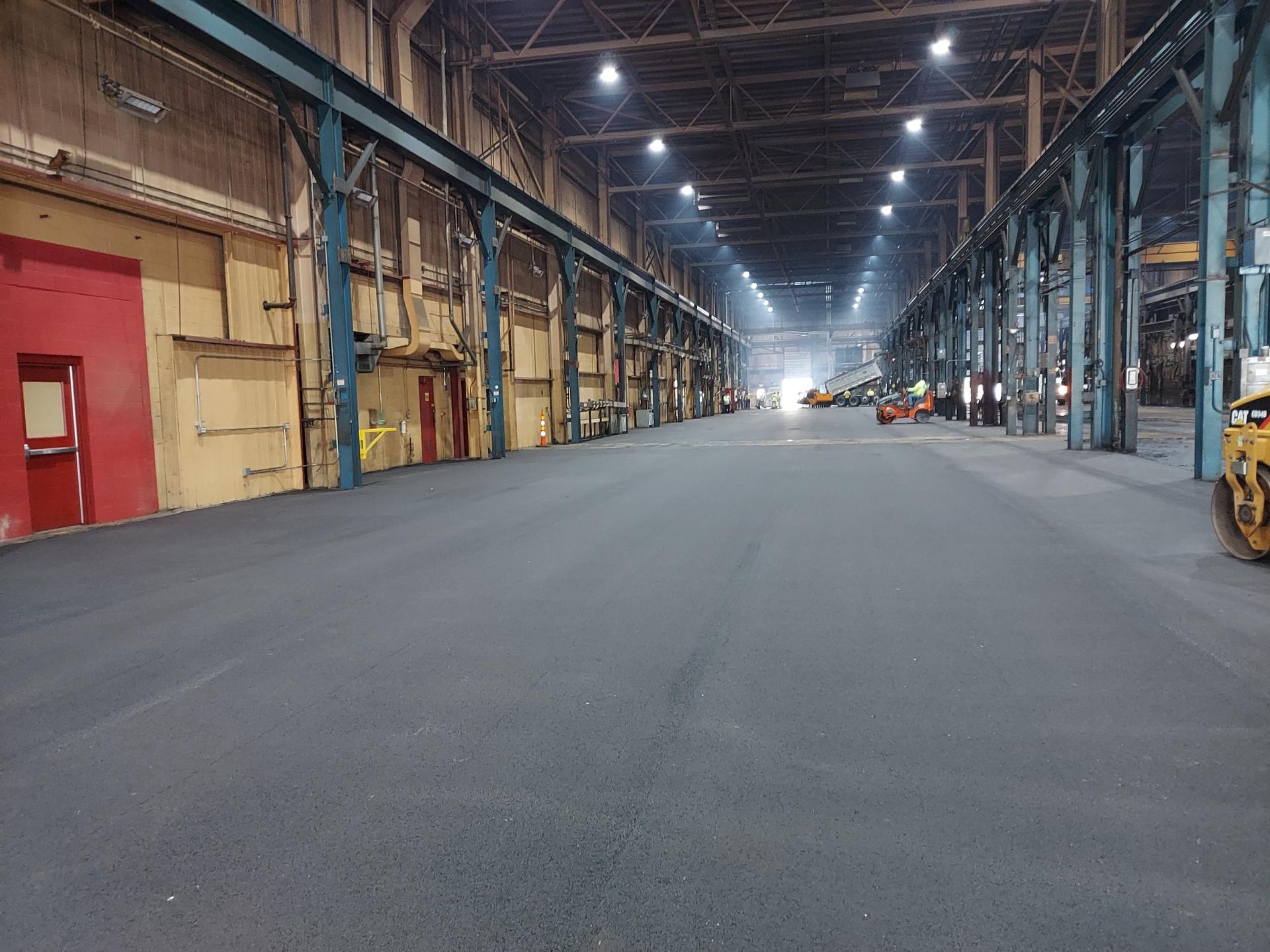 Workers use large rakes to spread freshly laid asphalt at busy construction site.