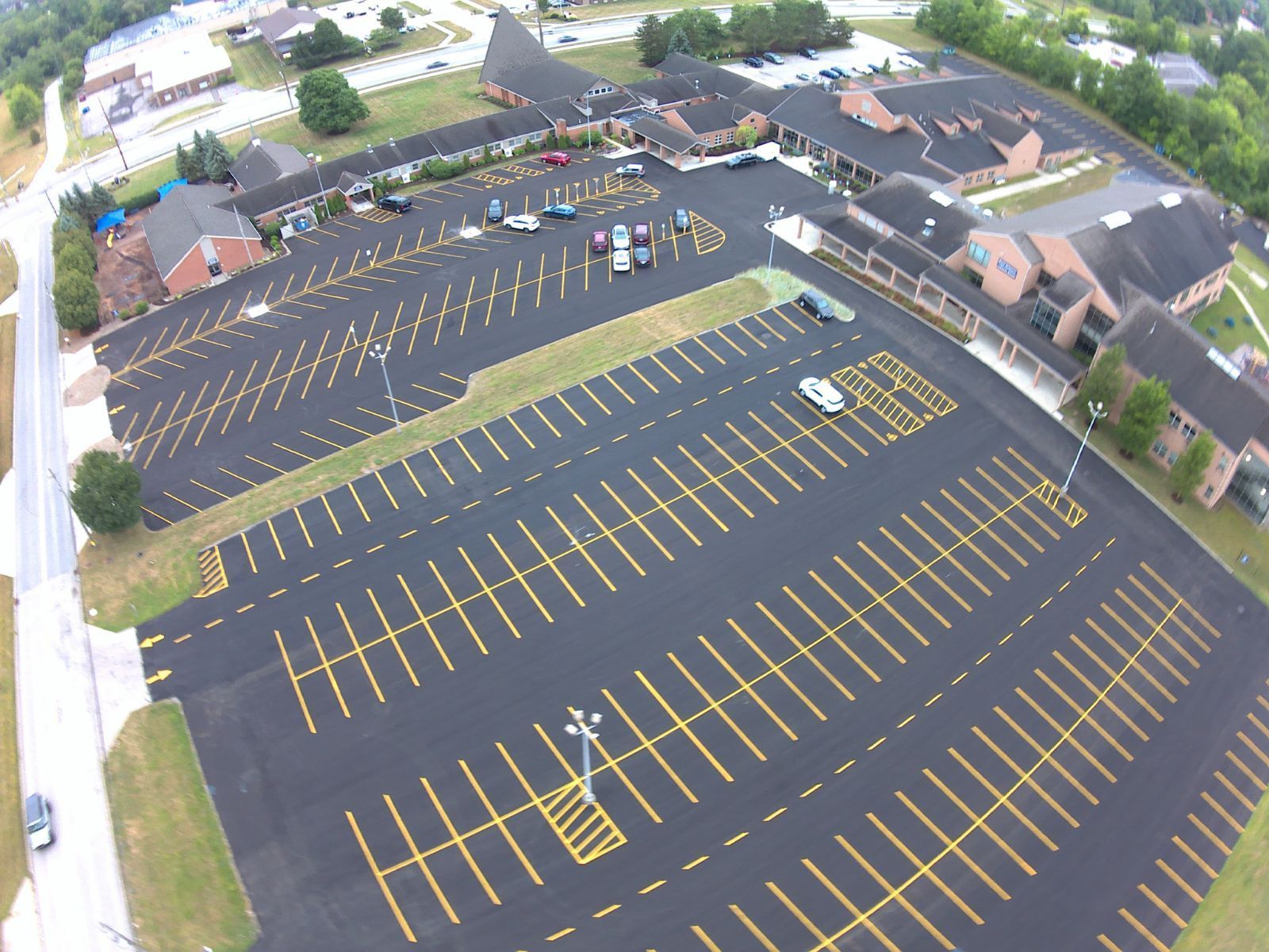 Aerial view of a mostly empty blacktop parking lot with bright yellow lines beside buildings and trees.